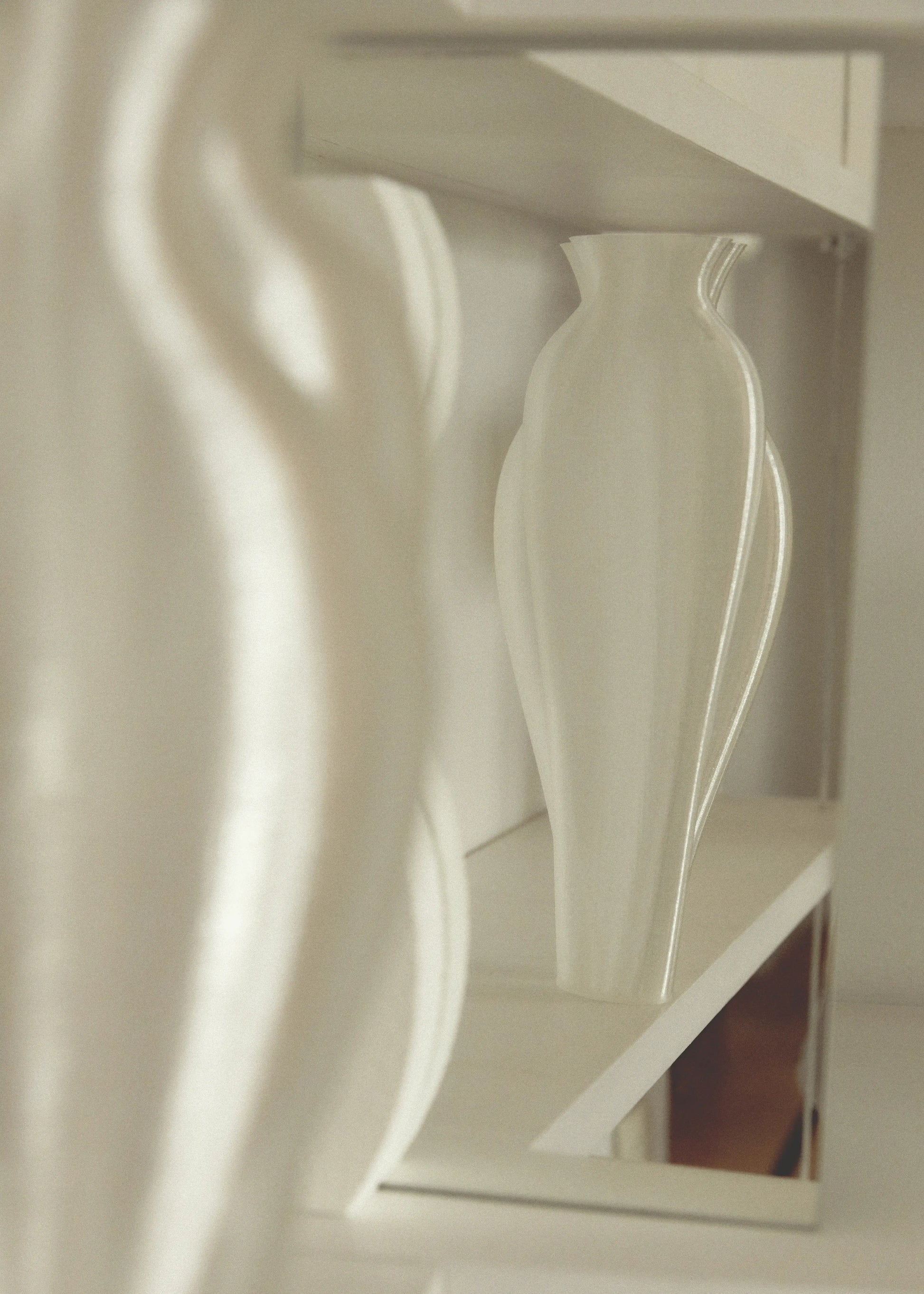 An Argot Studio Soeurs Vase with a curvy, modern design rests on a white shelf, its reflection in a nearby mirror creating symmetry. The scene is accented by soft, neutral tones.