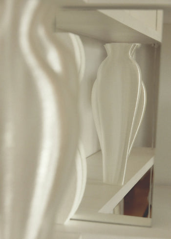 An Argot Studio Soeurs Vase with a curvy, modern design rests on a white shelf, its reflection in a nearby mirror creating symmetry. The scene is accented by soft, neutral tones.