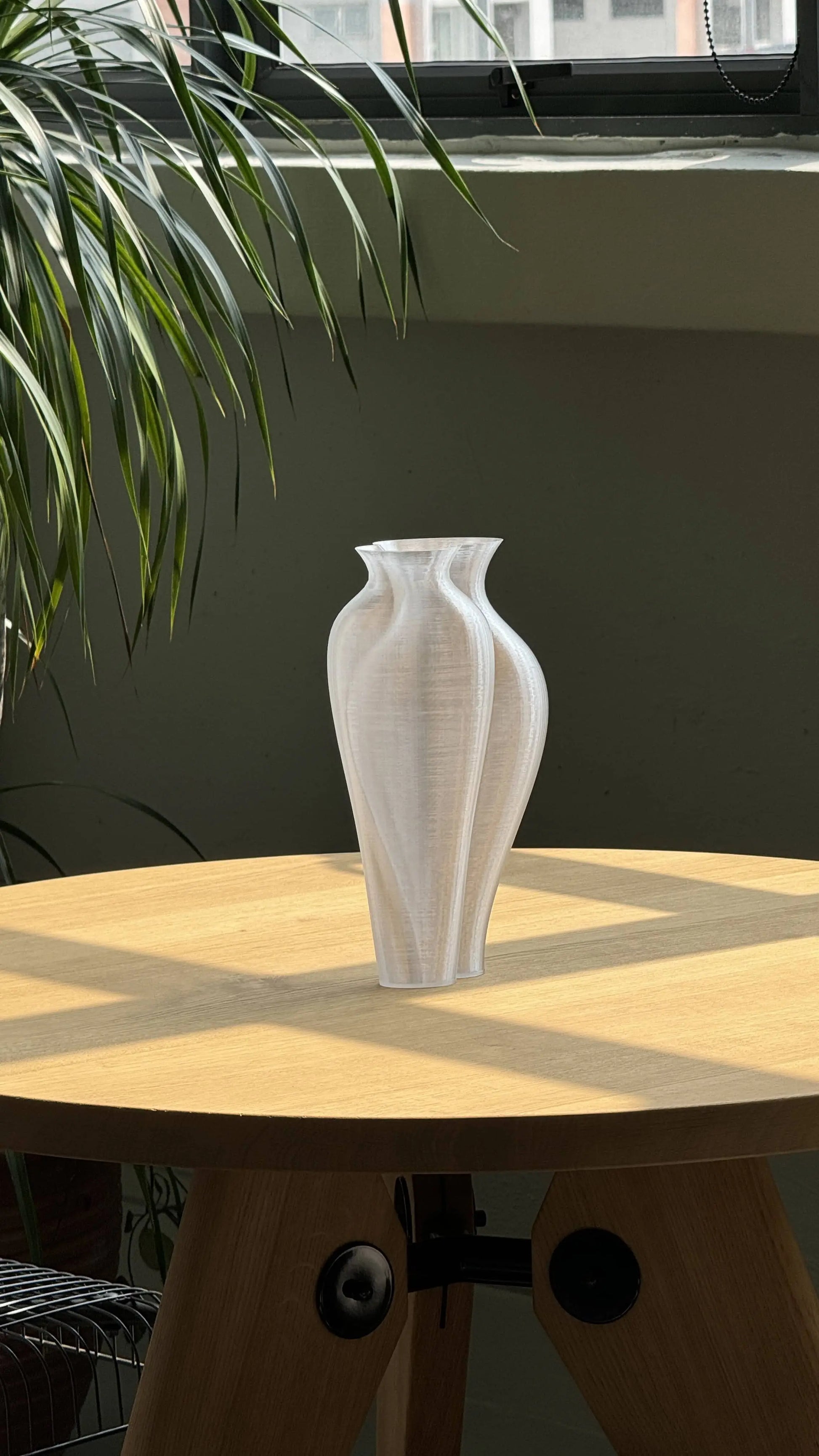 The Argot Studio Soeurs Vase, with its modern translucent design, rests on a sunlit round wooden table casting geometric shadows. Green plant leaves hang to the left and a window is visible in the background.
