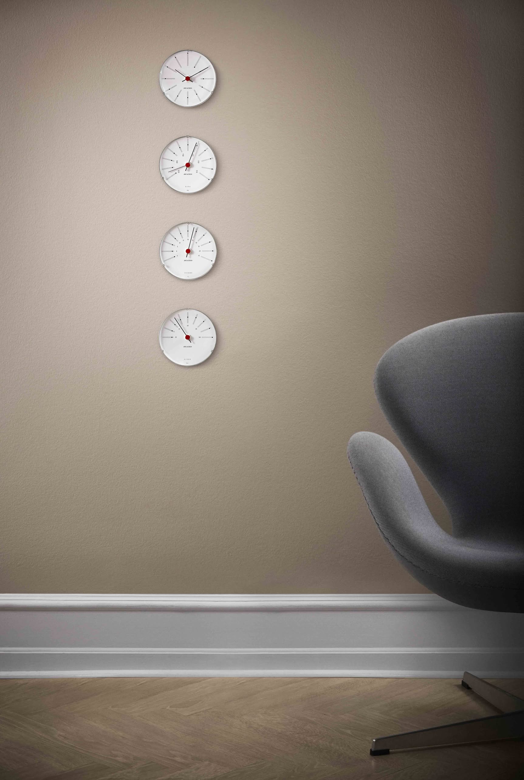 Four Arne Jacobsen Bankers Barometer 12cm pieces by Arne Jacobsen are mounted vertically on a beige wall, each displaying a different reading. A modern gray chair is partially visible at the lower right near the baseboard.