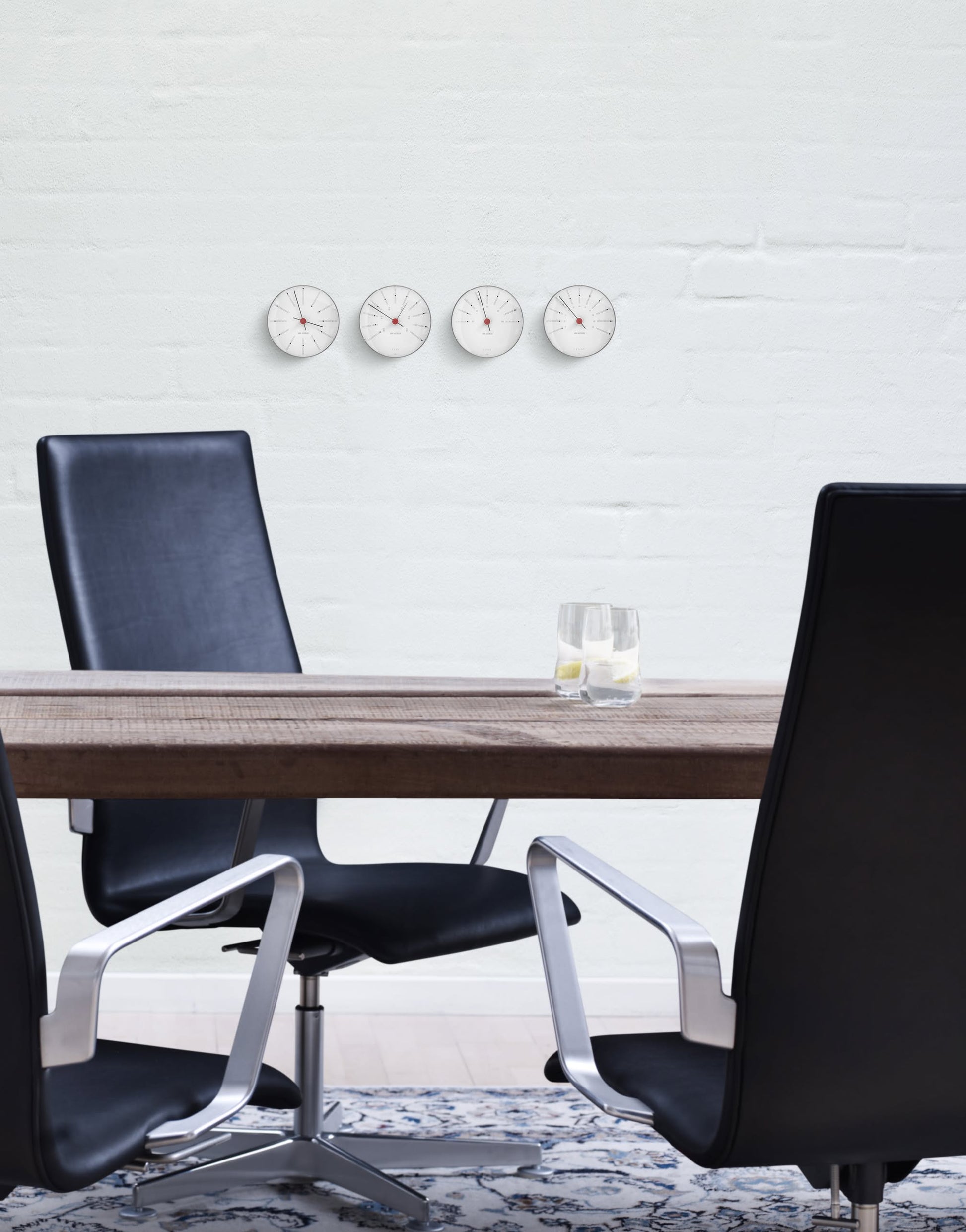 A modern conference room with minimalist elegance features a wooden table, two black chairs, a glass of lemon water, and four Arne Jacobsen Bankers Barometer 12cm clocks by Arne Jacobsen on a white wall showing different times.