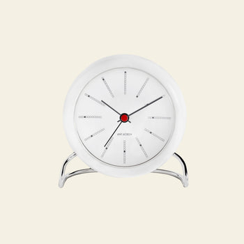 The Arne Jacobsen Bankers Table Clock 11cm by Arne Jacobsen is a modern white analog clock with a red center, black hands, and minimalist design on a silver stand, displayed against a light beige background.