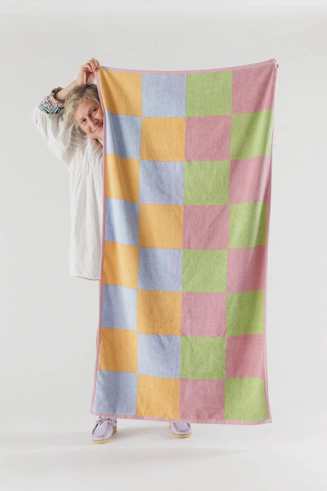 A person holds up the BAGGU Bath Towel, featuring a vibrant pink, yellow, green, and blue checkerboard design, partially concealing themselves against a plain white background. This striking towel is by BAGGU.