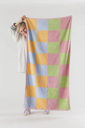 A person holds up the BAGGU Bath Towel, featuring a vibrant pink, yellow, green, and blue checkerboard design, partially concealing themselves against a plain white background. This striking towel is by BAGGU.