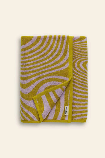 A folded BAGGU Bath Towel by BAGGU, featuring a vibrant wavy mustard yellow and lavender abstract print with a small white brand label on the edge, set against a plain light beige background—ideal as a sunflower-inspired bath towel accent.