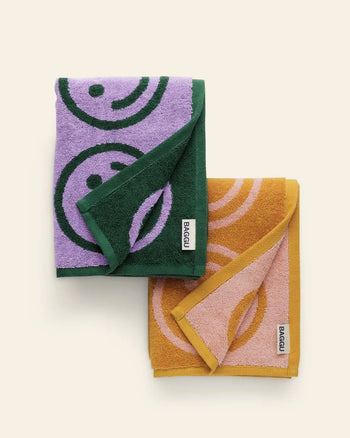 The BAGGU Hand Towel Set of 2 features cheerful smiley face designs—one in purple with green, the other in mustard yellow and pink—each with a white BAGGU label, perfect for brightening any bathroom cleanup.