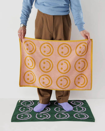 A person stands on a green mat with purple smiley faces, holding the BAGGU Hand Towel Set of 2. Both towels feature the brands cheerful smiley face design, making cleanup feel bright and fun.