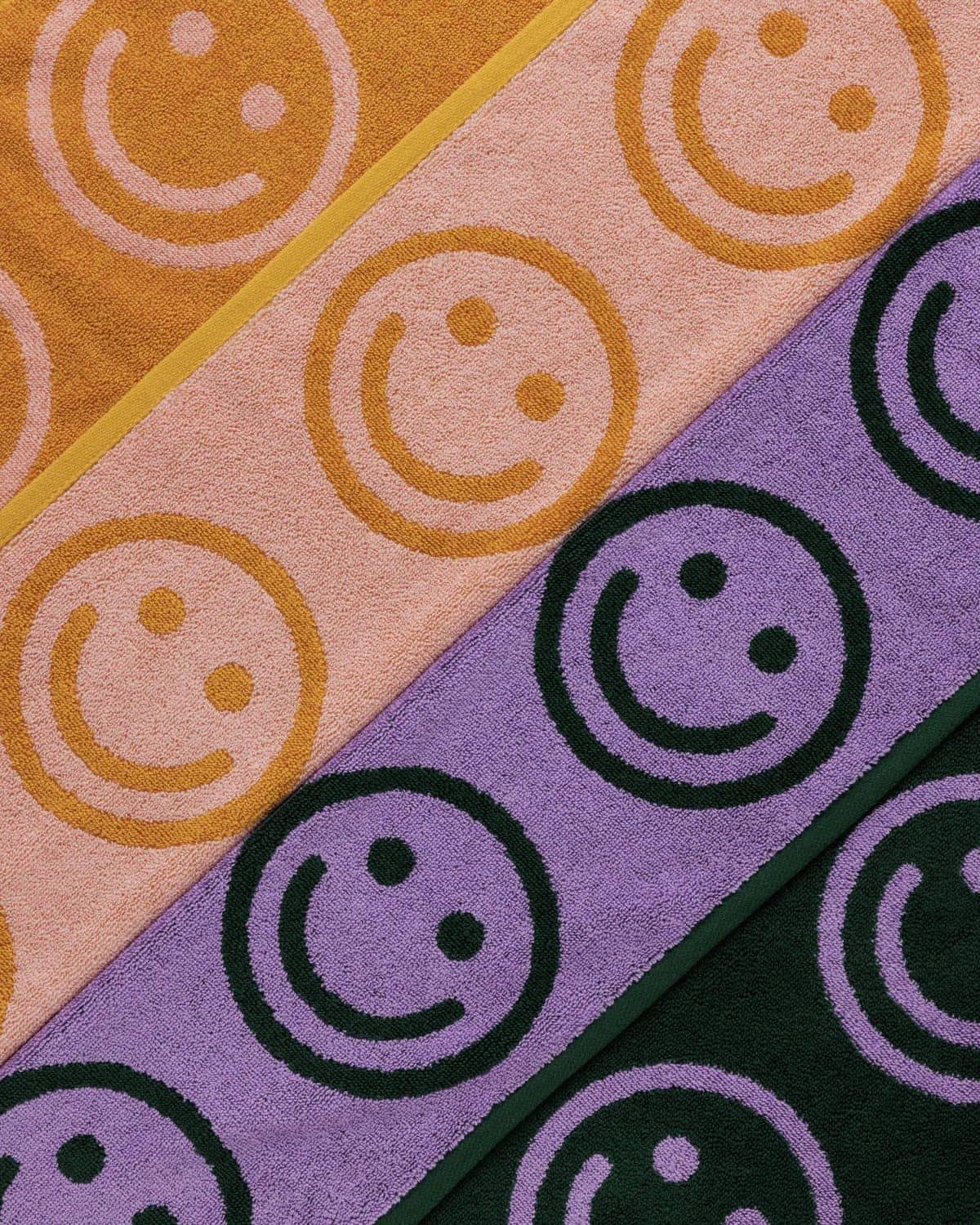 Diagonal rows of BAGGU Hand Towel Set of 2 by BAGGU, shown in yellow, pink, purple, and dark green. Each towel features a bold smiley face design for a cheerful cleanup.