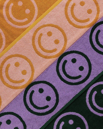 Diagonal rows of BAGGU Hand Towel Set of 2 by BAGGU, shown in yellow, pink, purple, and dark green. Each towel features a bold smiley face design for a cheerful cleanup.