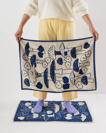 Wearing yellow pants and a white sweater, someone holds a BAGGU Hand Towel Set of 2 with a mushroom and flower print from BAGGU, standing on a matching blue mat and wearing purple socks—a cheerful touch for cleanup with this smiley face design.