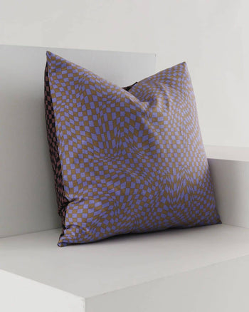 The BAGGU Throw Pillow Case by BAGGU showcases a trippy purple and brown checker design, adding flair to any living space as it rests against a white minimalist bench and wall.