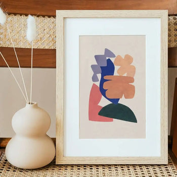 The Bbblob Floating In Mid Air, 2020 - Giclee Print (21x30) by Bbblob stands framed on a woven surface beside a matte beige vase with three dried white grasses, set against warm wooden tones for a peaceful ambiance.