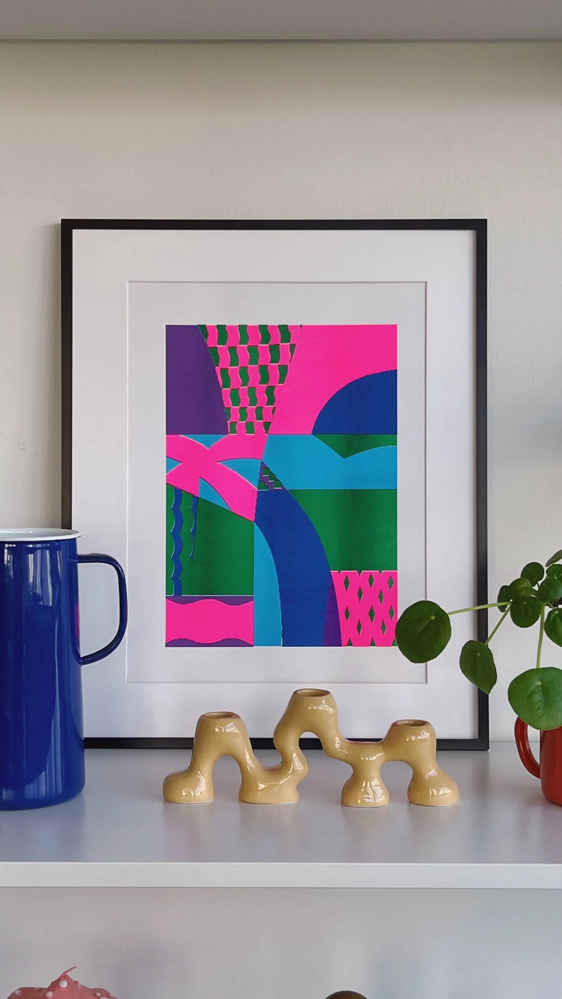 Bbblob Freefall, 2022, a Risograph print (30x42) in bold pink, green, and blue shapes in a black frame. Displayed on a shelf with a blue mug, beige ceramic sculpture, and red potted plant. Playful misregistration adds character.
