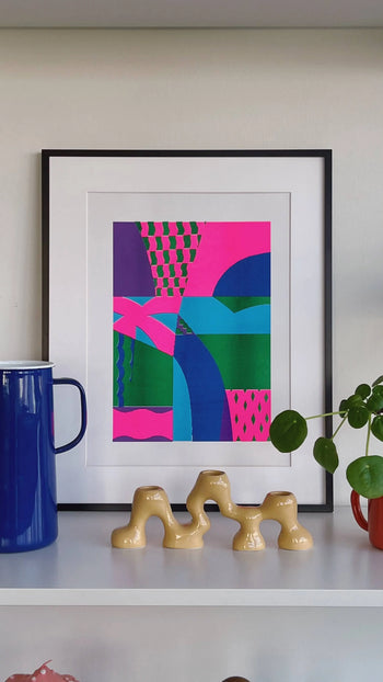 Bbblob Freefall, 2022, a Risograph print (30x42) in bold pink, green, and blue shapes in a black frame. Displayed on a shelf with a blue mug, beige ceramic sculpture, and red potted plant. Playful misregistration adds character.