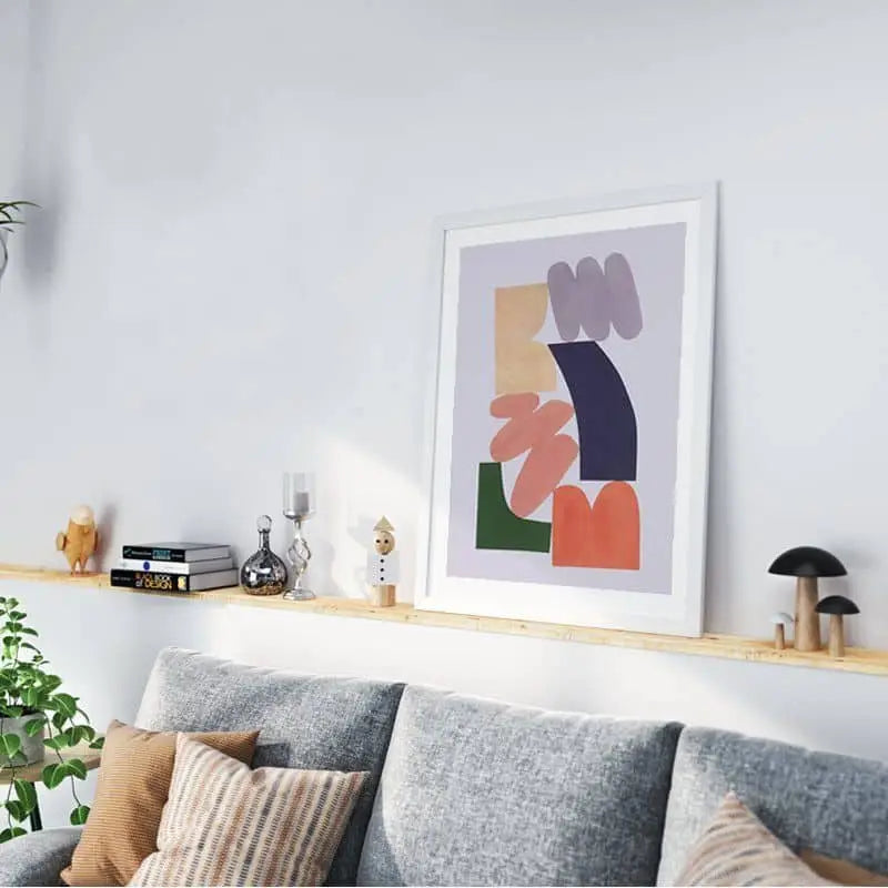A modern living room with a gray sofa, patterned cushions, wooden shelf with books and decor, and Bbblob Lift You Up, 2020 Giclee Print (30x40) on the white wall. A green plant in the corner adds freshness to the space.