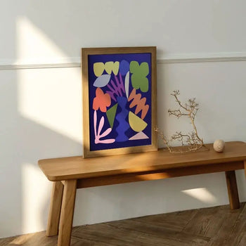 The Bbblob Morph, 2020 - Fine Art Print (32x44) by Bbblob features vibrant abstract shapes. Framed and displayed on a wooden bench, its joined by a bare branch and round decor as sunlight casts dynamic shadows against a light wall.