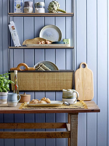 A rustic mahogany wood table with mugs, a teapot, and pastries sits before a shelf displaying plates, cups, books, a loaf of bread, and the Bloomingville Abbas Cutting Board. A basil plant rests on the table next to the crockery.