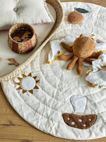 A soft Bloomingville Agnes Blanket from Bloomingville with stitched sun and spaceship designs rests on the wooden floor, accompanied by a plush octopus toy, a woven basket filled with small wooden toys, and a patterned pillow.