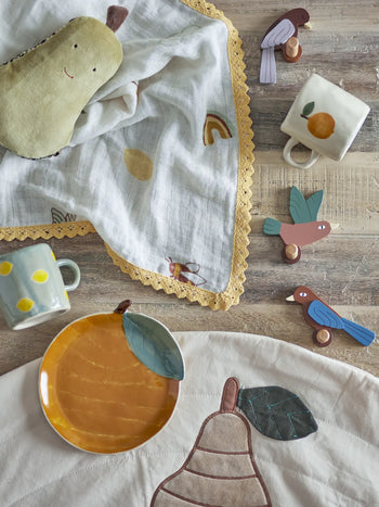 A flat lay of baby items on wood, including a pear plush, the Bloomingville Agnes Blanket, two ceramic mugs, wooden bird toys, a yellow pear plate, and a white cloth with a pear applique by Bloomingville MINI.