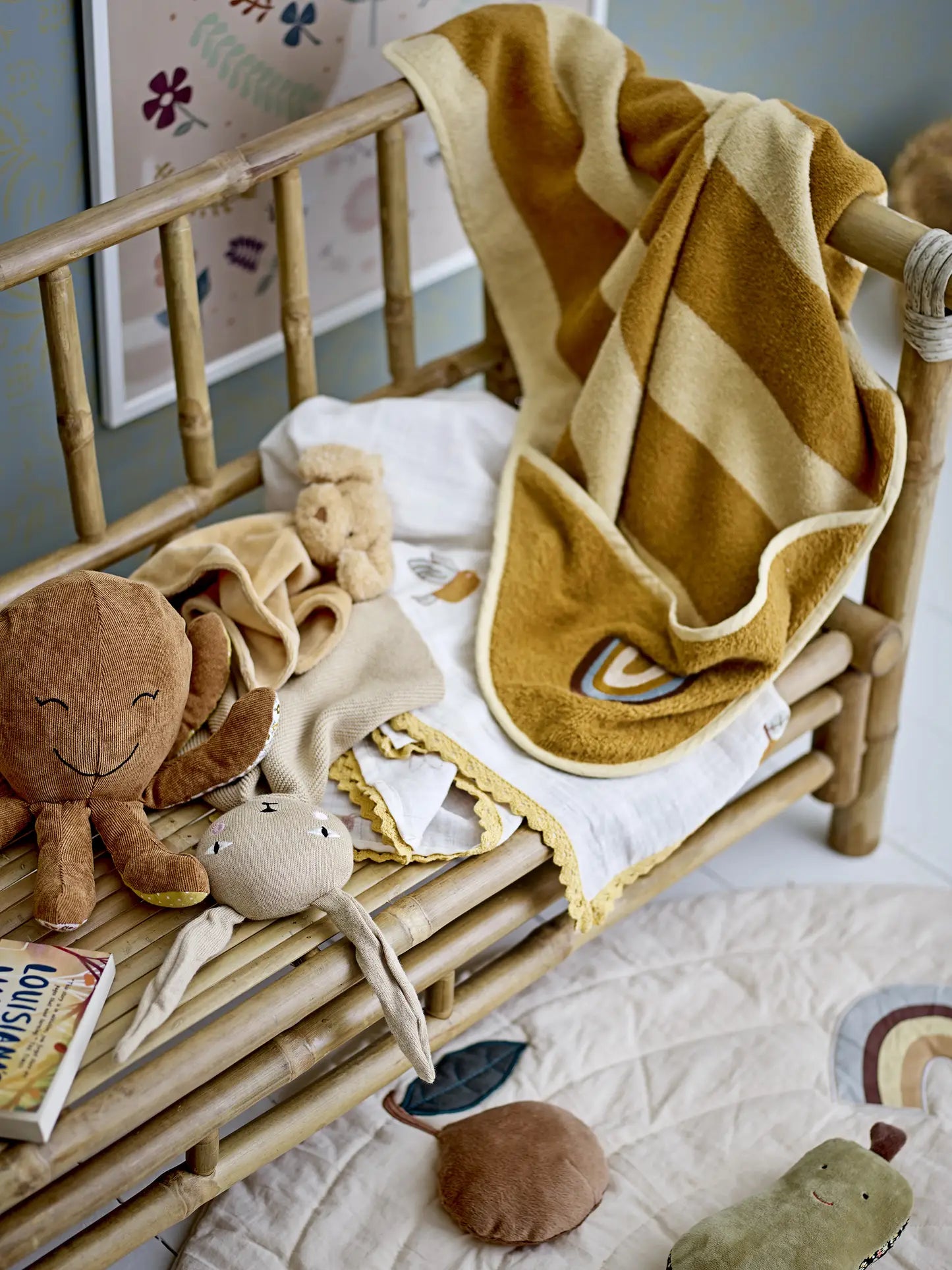 A wooden crib holds a Bloomingville Agnes Blanket in mustard-yellow stripes, soft toys (monkey, rabbit, and bear), and a rainbow-print cloth. A Bloomingville MINI quilted play mat with toys is on the floor nearby.