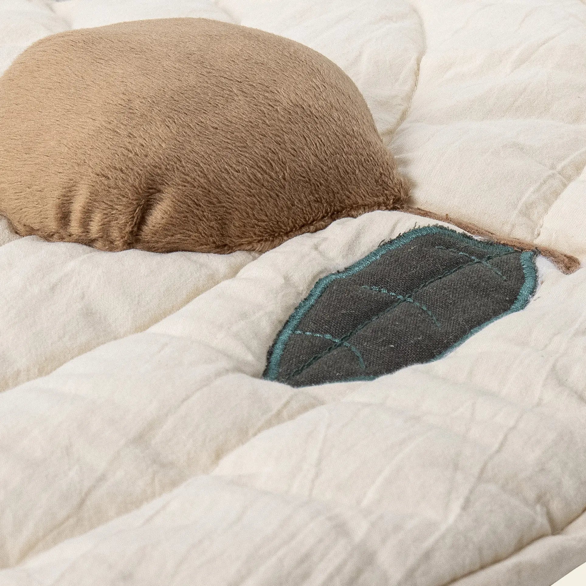 A close-up of the Bloomingville Agnes Play Mat by Bloomingville, showcasing its beige quilted design with a soft, round brown pillow and a leaf-shaped patch with blue stitching for gentle sensory exploration.