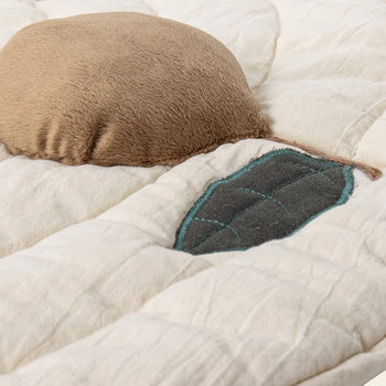 A close-up of the Bloomingville Agnes Play Mat by Bloomingville, showcasing its beige quilted design with a soft, round brown pillow and a leaf-shaped patch with blue stitching for gentle sensory exploration.