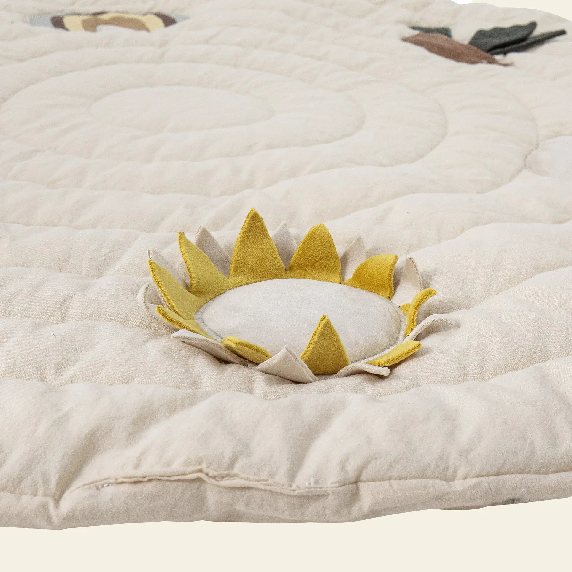 The Bloomingville Agnes Play Mat by Bloomingville is a soft, cream-colored quilt with a fabric sunflower—yellow petals and white center—plus stitched spirals and small sensory details, ideal for tactile exploration.