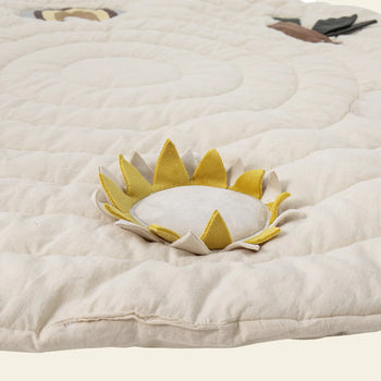 The Bloomingville Agnes Play Mat by Bloomingville is a soft, cream-colored quilt with a fabric sunflower—yellow petals and white center—plus stitched spirals and small sensory details, ideal for tactile exploration.