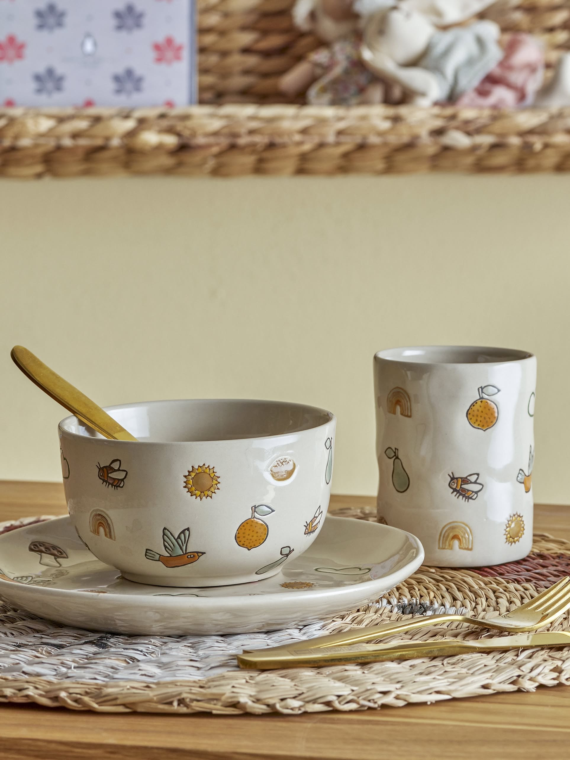 The Bloomingville Agnes Tableware Set by Bloomingville features a handmade ceramic bowl with yellow spoon, matching plate and cup, all with whimsical fruit, rainbow, and sun motifs on a woven placemat for unique charm.