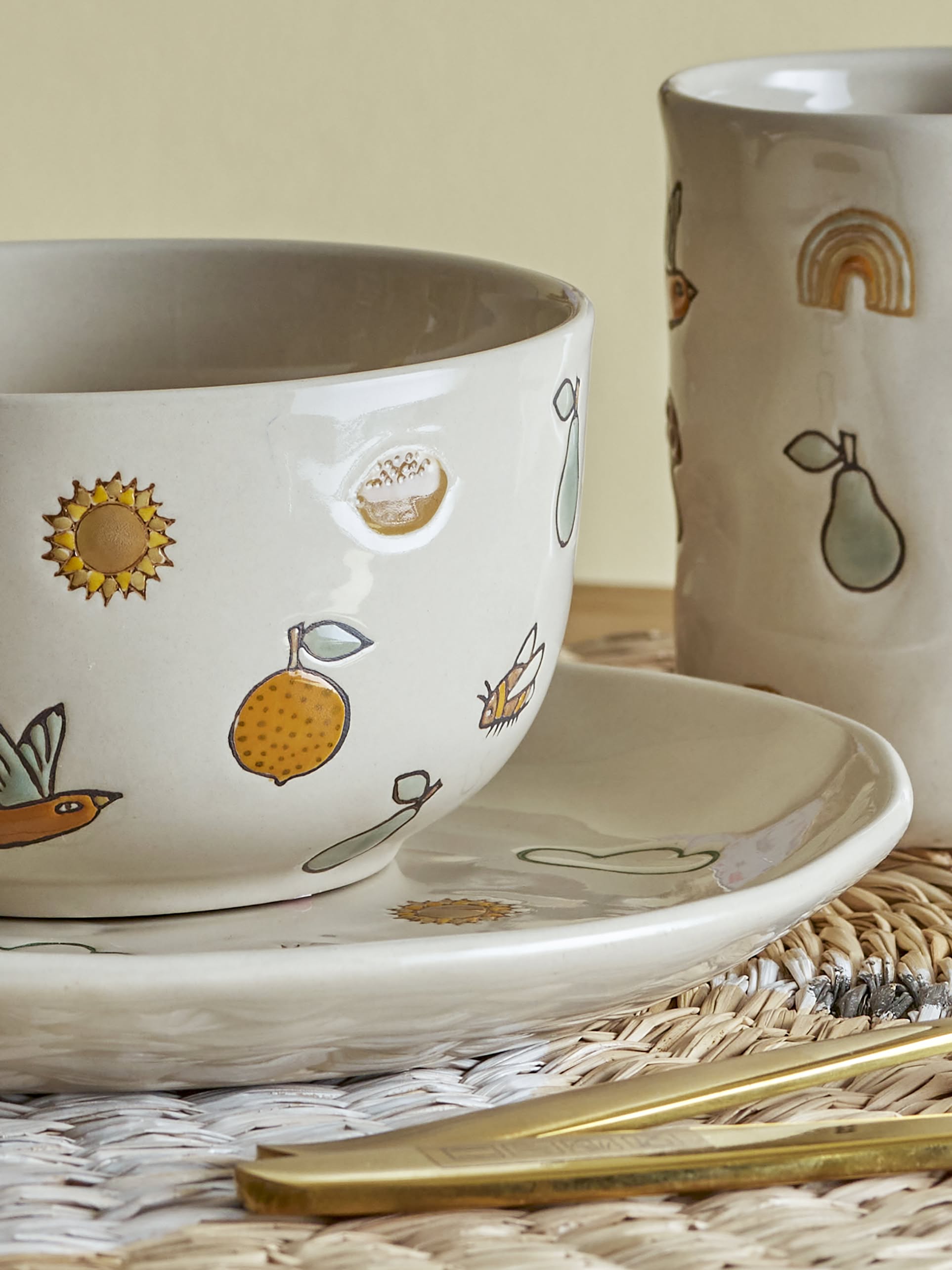 The Bloomingville Agnes Tableware Set by Bloomingville features a handmade ceramic cup, mug, and plate decorated with lemons, pears, a sunflower, and a rainbow—each piece brimming with unique charm on a woven placemat with gold cutlery.