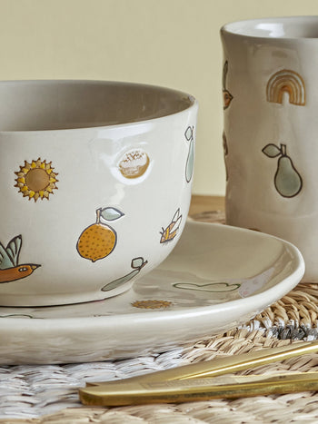 The Bloomingville Agnes Tableware Set by Bloomingville features a handmade ceramic cup, mug, and plate decorated with lemons, pears, a sunflower, and a rainbow—each piece brimming with unique charm on a woven placemat with gold cutlery.