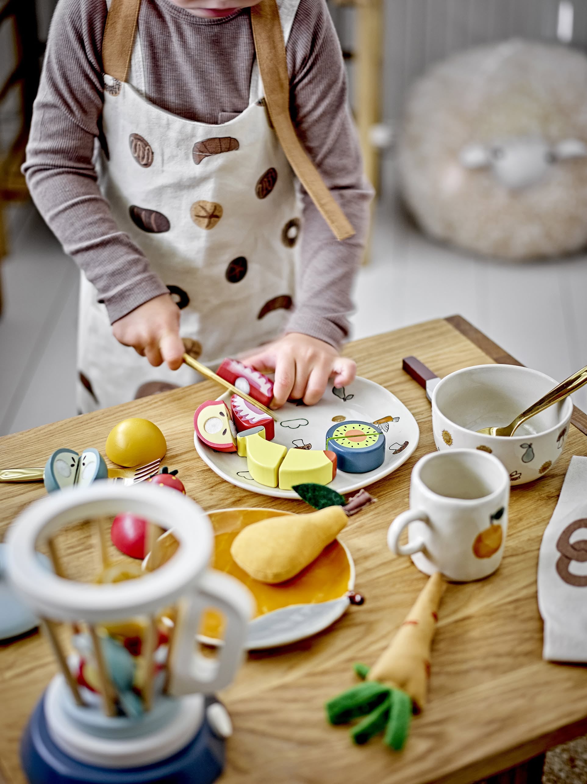 A child in an apron plays with the Bloomingville Agnes Tableware Set, using a play knife to cut colorful wooden toy fruits and vegetables on a plate at a small table set for charming pretend play.