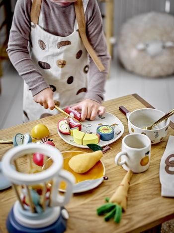 A child in an apron plays with the Bloomingville Agnes Tableware Set, using a play knife to cut colorful wooden toy fruits and vegetables on a plate at a small table set for charming pretend play.