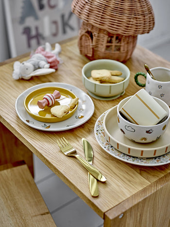 A wooden table set with the Bloomingville Agnes Tableware Set, heart and star-shaped cookies, gold-colored cutlery, a toy bunny, and a wicker house in the background creates a playful, cozy scene full of unique charm.