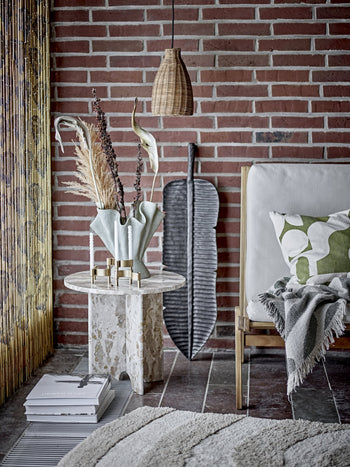 A cozy living space with a brick wall, a white cushioned chair, a green and white pillow, and the Bloomingville Alk Throw in recycled cotton. A marble side table with decor sits under a pendant lamp.