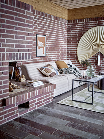 A cozy living room with red brick walls, a beige sofa topped with the Bloomingville Alk Throw, minimalist cushions, a modern coffee table, vases, artwork, and fan-shaped wall decor. Tiled floors and warm hues make the space inviting.