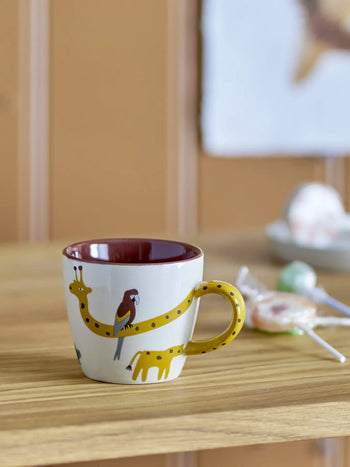 A Bloomingville Belina Cup by Bloomingville, featuring animal motifs—a giraffe, bird, and elephant—yellow handle, and maroon interior, sits on a wooden surface with lollipops in the foreground and a blurred background.