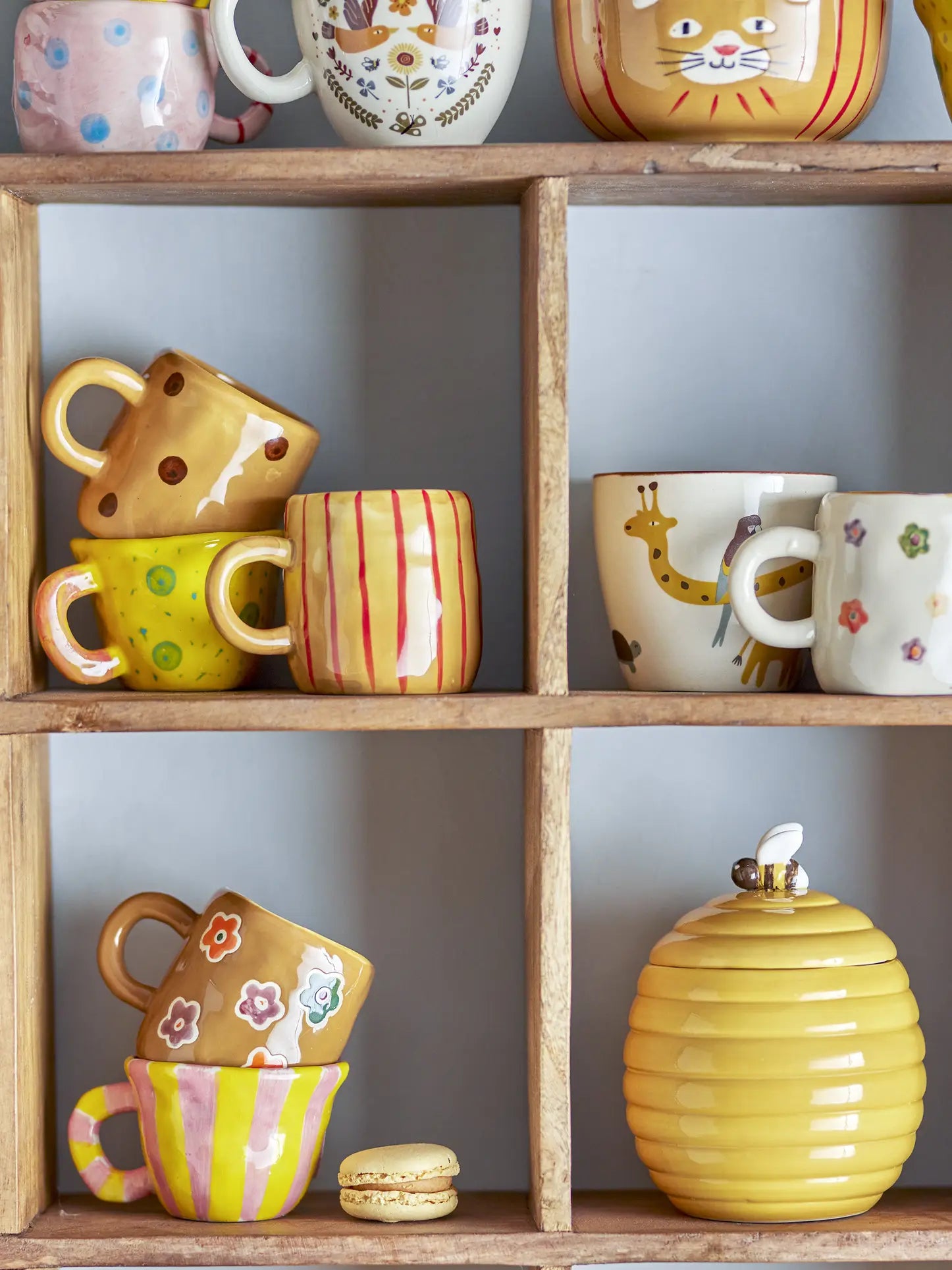 A wooden shelf displays colorful ceramic mugs and the Bloomingville Belina Cup, featuring playful patterns like dots, stripes, flowers, and animal motifs. A yellow beehive-shaped jar with a lid and a yellow macaron rest on the bottom shelf.