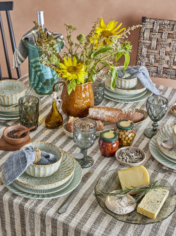 A cozy meal scene features a rustic table set with striped woven cotton linens, ceramic plates and bowls, vintage glasses, cheese, bread, olives, preserves, sunflowers in a pitcher—and the Bloomingville Binnur Throw by Bloomingville adds inviting texture.