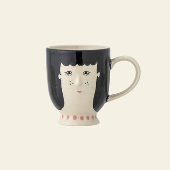 The Bloomingville Carolin Cup by Bloomingville is a ceramic mug with a handle, hand-painted with a whimsical face—black hair, closed eyes, freckles, red lips—and features a pink dotted base. The background is plain light cream.