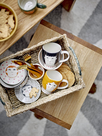 A woven basket holds plates, Bloomingville Carolin Cups featuring hand-painted faces, a sheep-adorned tin, gold-colored cutlery, and an animal-shaped cookie. The basket rests on a wooden stool beside snacks and above a geometric-patterned rug.