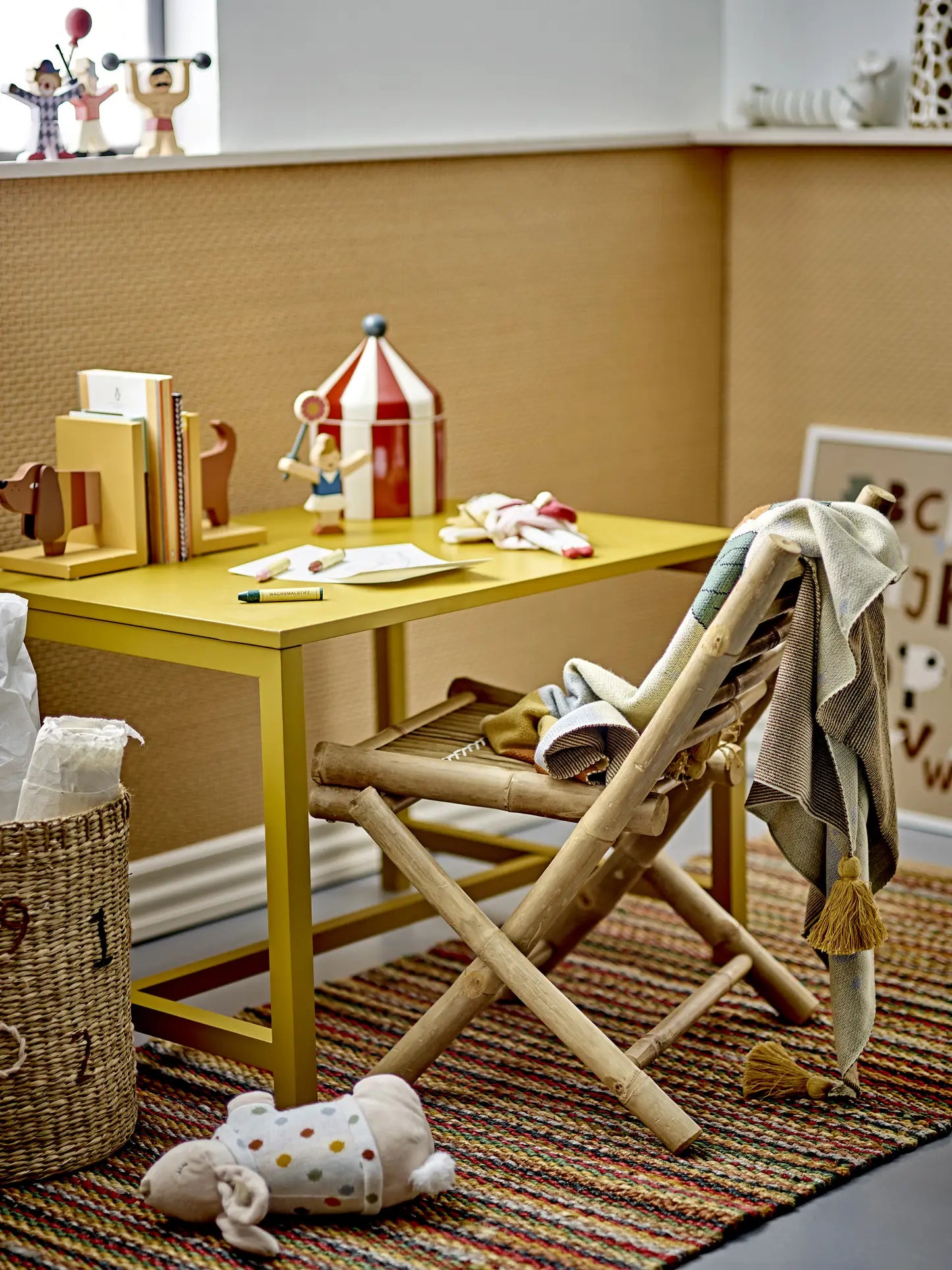 A small yellow desk with children’s books, toys, and colored pencils sits by a wooden chair with a blanket. The Bloomingville Charlie Bookend brings charm to the children’s room, while a plush toy rests on the striped rug nearby.