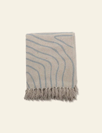 The Bloomingville Dami Throw by Bloomingville is a folded soft blanket made from recycled cotton, featuring subtle wavy blue lines and fringed tassels on one edge, displayed on a light background for sustainable style.