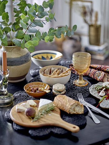 A cozy, inviting kitchen scene is set with a stylish table featuring a vase of eucalyptus, candle, patterned snack dishes, glass goblet, and cheese and bread on the Bloomingville Dana Cutting Board by Bloomingville.