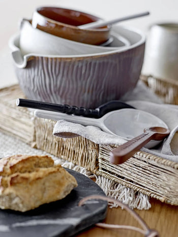 A stack of rustic ceramic bowls and spoons rests on a textured placemat next to crusty bread atop the Bloomingville Daris Cutting Board, adding a cozy, artisanal touch to the table.