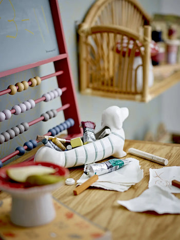 A wooden table holds art supplies—paint tubes in the Bloomingville Djaco Bowl, apples on a plate, chalk, and paper. An abacus and shelves with decorative jars from Bloomingville create a cozy, creative workspace in the background.
