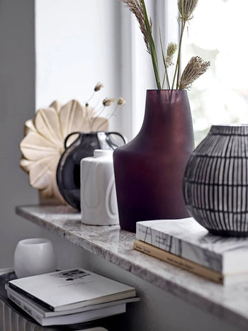 A marble shelf displays decorative vases, like the deep purple Bloomingville Elice Vase, some filled with dried grasses. Neatly stacked books and natural light combine to create a fresh, inviting home atmosphere.