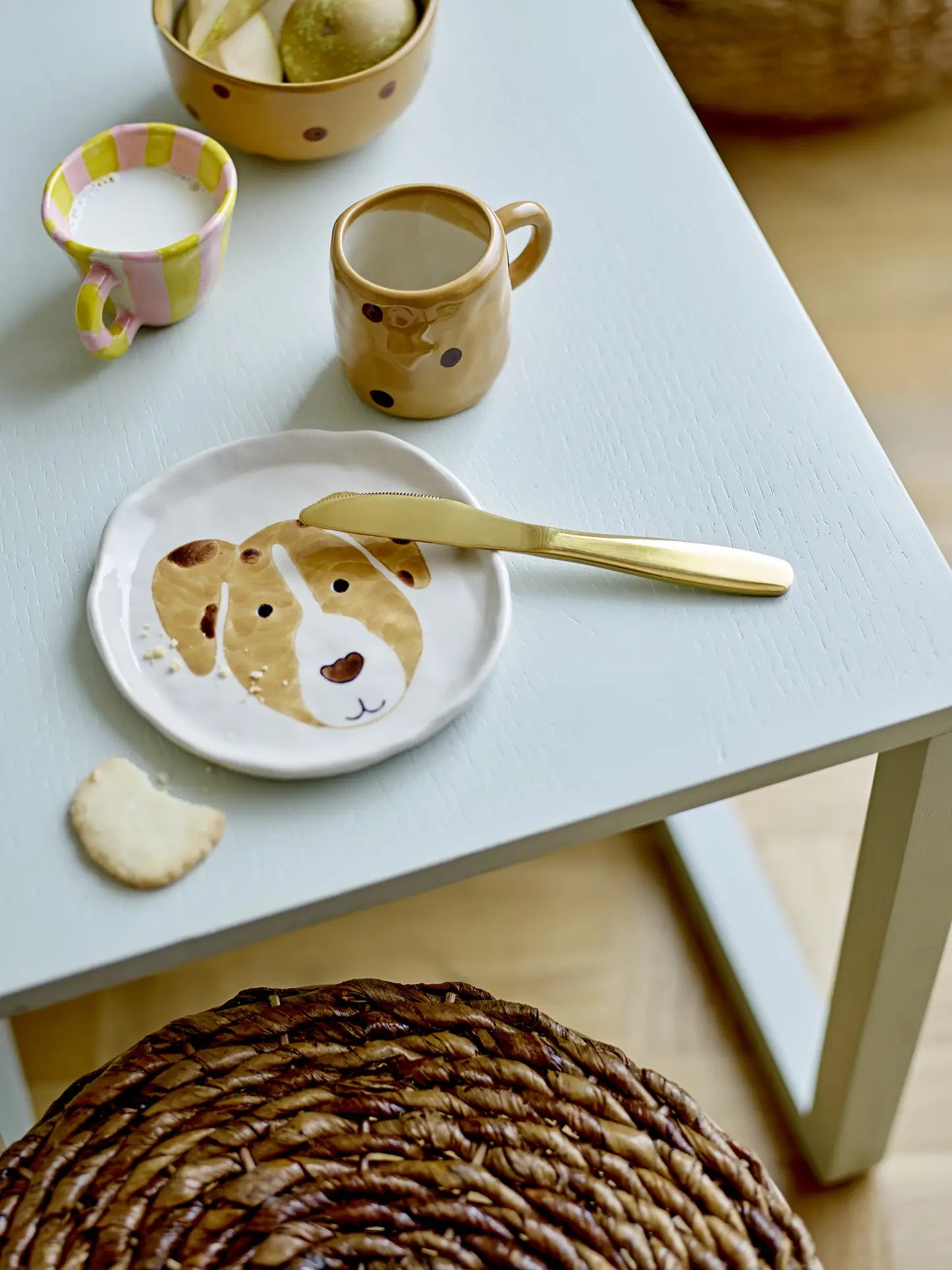 A white table is set with a Bloomingville Fenix Tableware plate featuring a dog motif, gold knife, cup of milk, dotted brown Bloomingville mug, bowl of fruit, heart-shaped cookie, and has a woven stool nearby.