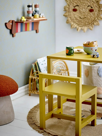 A cozy childrens playroom with a yellow table and chair, playful mugs and bowls from Bloomingville Fenix Tableware, a toy car, woven rug, wall art, a mushroom-shaped stool, and a colorful dog wall shelf with art supplies.