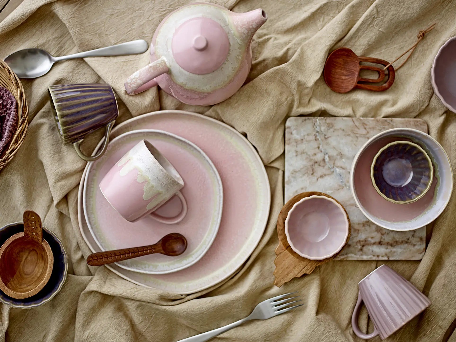 A flat lay of pink ceramic tableware and wooden spoons rests on a beige cloth, featuring the Bloomingville Gabrielle Trivet by Bloomingville for table protection. The pastel pieces have a handmade look, complemented by a fork and small basket.